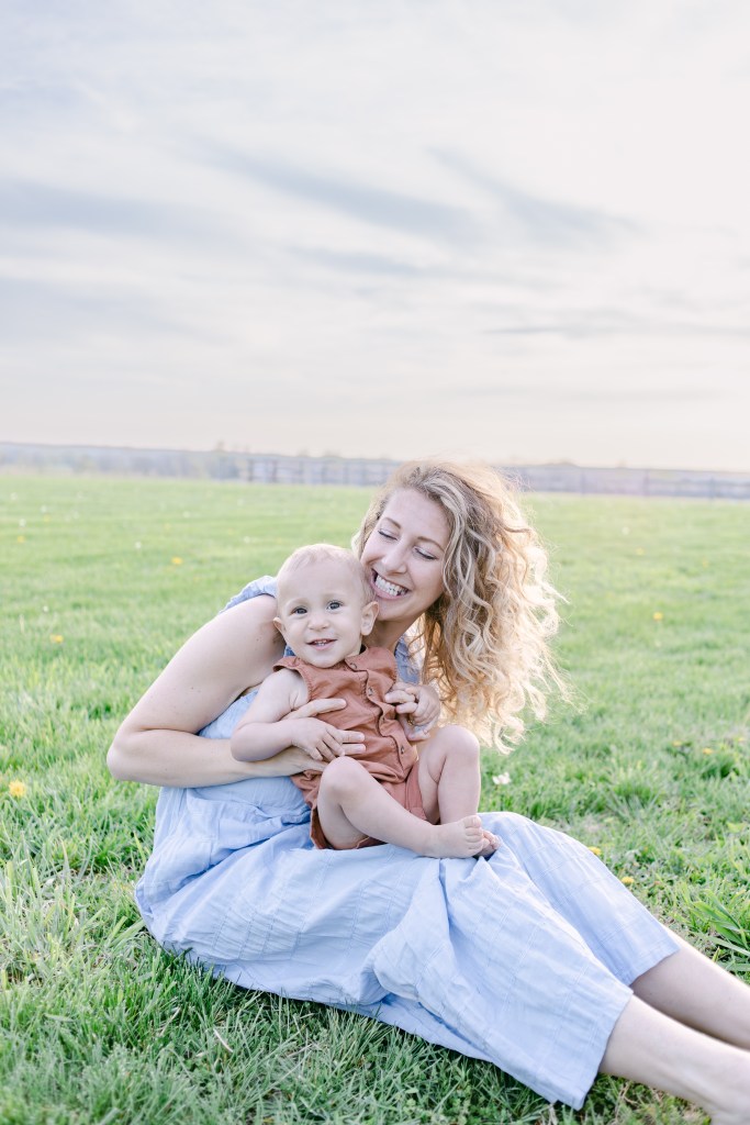 young mother smiles while holding young son in her lap while posting in green field 