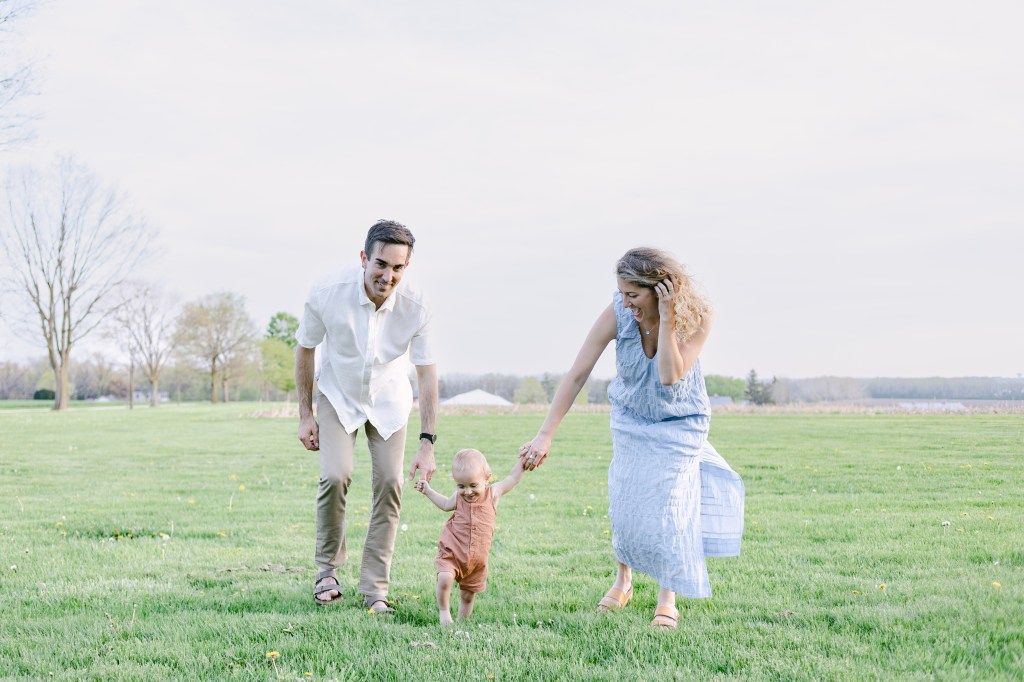 family of three holds hands while walking through green field, smiling 